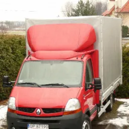 Czerwony samochód dostawczy Renault z plandeką w pochmurny dzień, widoczny numer rejestracyjny OP 87190, zarys zabudowań mieszkalnych w tle.